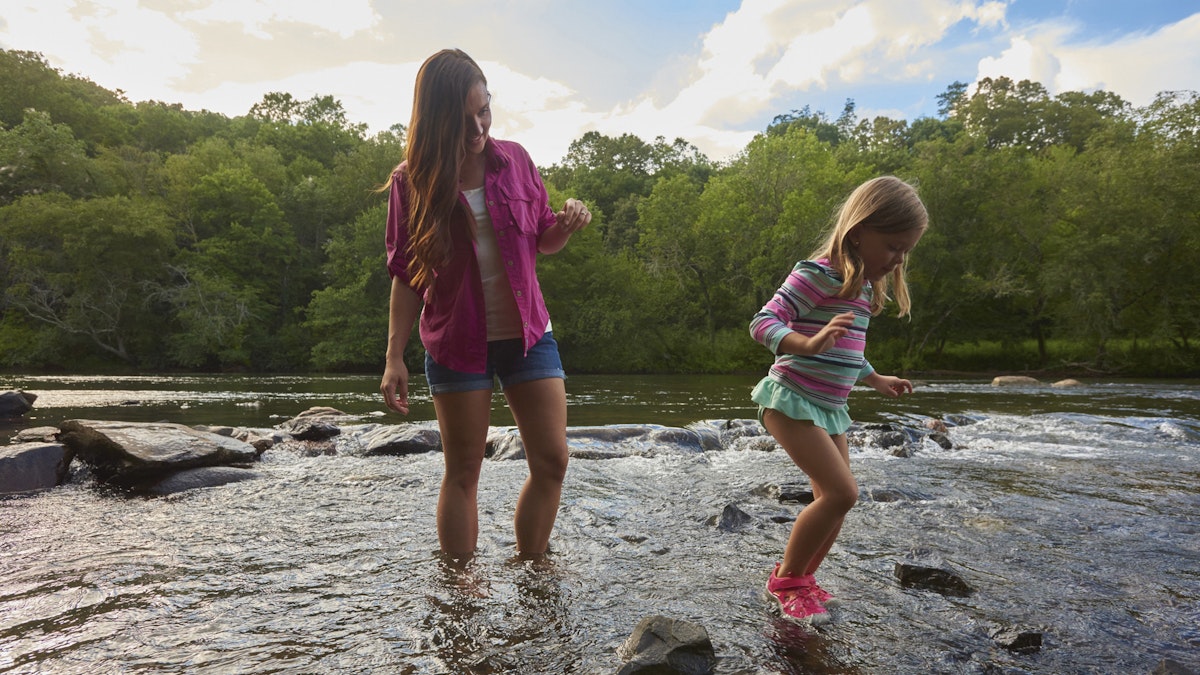A mother and daughter play in the shallow waters of a river. There are lush green trees lining the river in the distance.