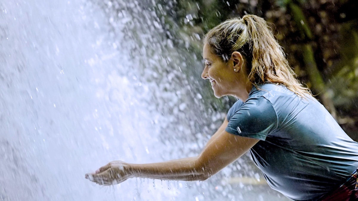 A woman leans forwards and cups her hands under a waterfall.
